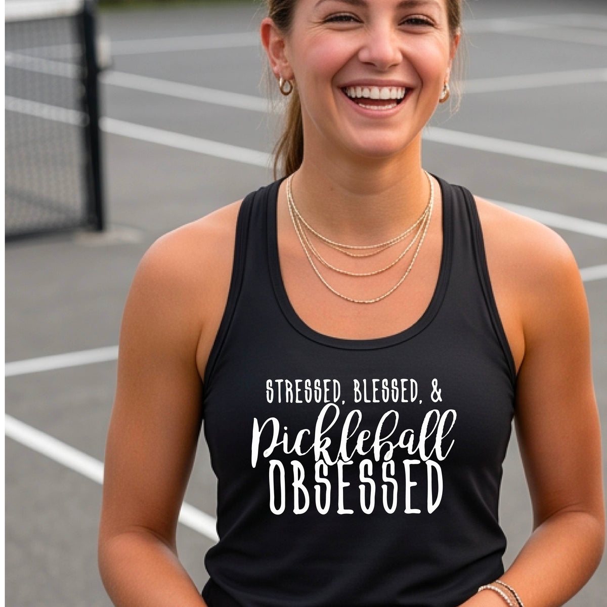 A woman on a pickleball court wearing a black racer-back tank top with a white graphic of the words “Stressed, Blessed & Pickleball Obsessed” on the front of the shirt.