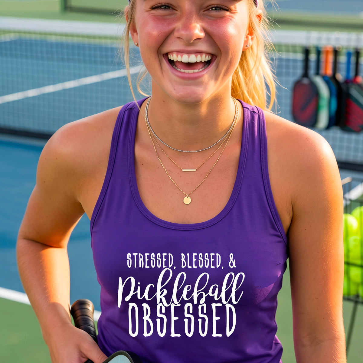 A woman on a pickleball court wearing a purple racerback tank top with a white graphic of the words “Stressed, Blessed & Pickleball Obsessed” on the front of the shirt.