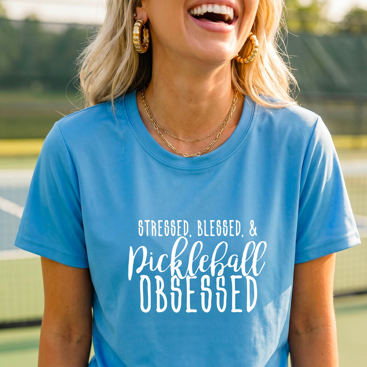 A woman on a pickleball court wearing a light blue short-sleeve t-shirt with a white graphic of the words “Stressed, Blessed & Pickleball Obsessed” on the front of the shirt.