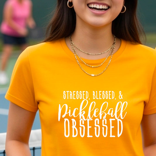 A woman on a pickleball court wearing a mustard yellow gold short-sleeve t-shirt with a white graphic of the words “Stressed, Blessed & Pickleball Obsessed” on the front of the shirt.
