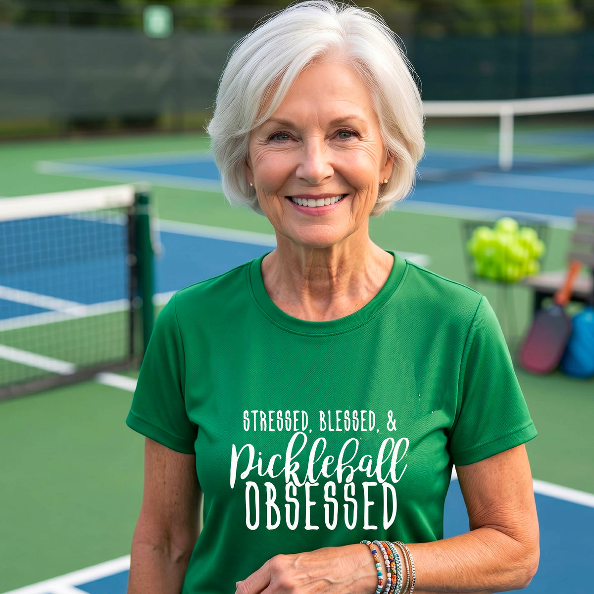 A woman on a pickleball court wearing a green short-sleeve t-shirt with a white graphic of the words “Stressed, Blessed & Pickleball Obsessed” on the front of the shirt.