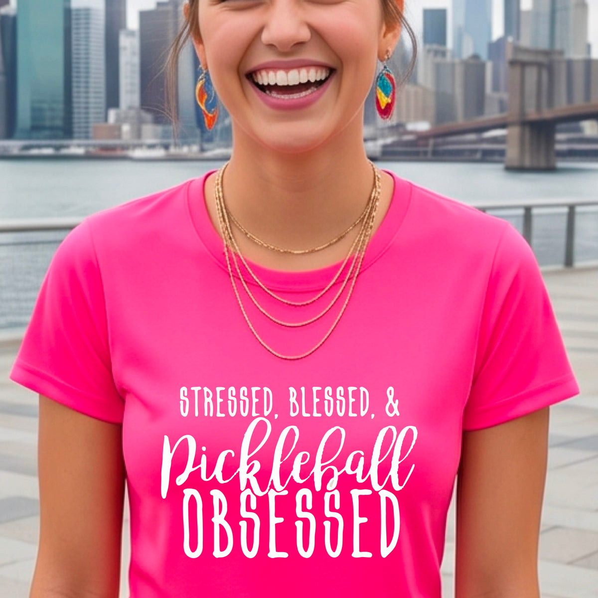 A woman wearing a pink short-sleeve t-shirt with a white graphic of the words “Stressed, Blessed & Pickleball Obsessed” on the front of the shirt.