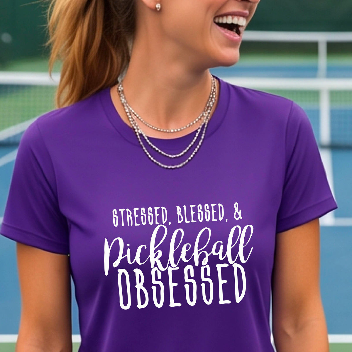 A woman on a pickleball court wearing a purple short-sleeve t-shirt with a white graphic of the words “Stressed, Blessed & Pickleball Obsessed” on the front of the shirt.