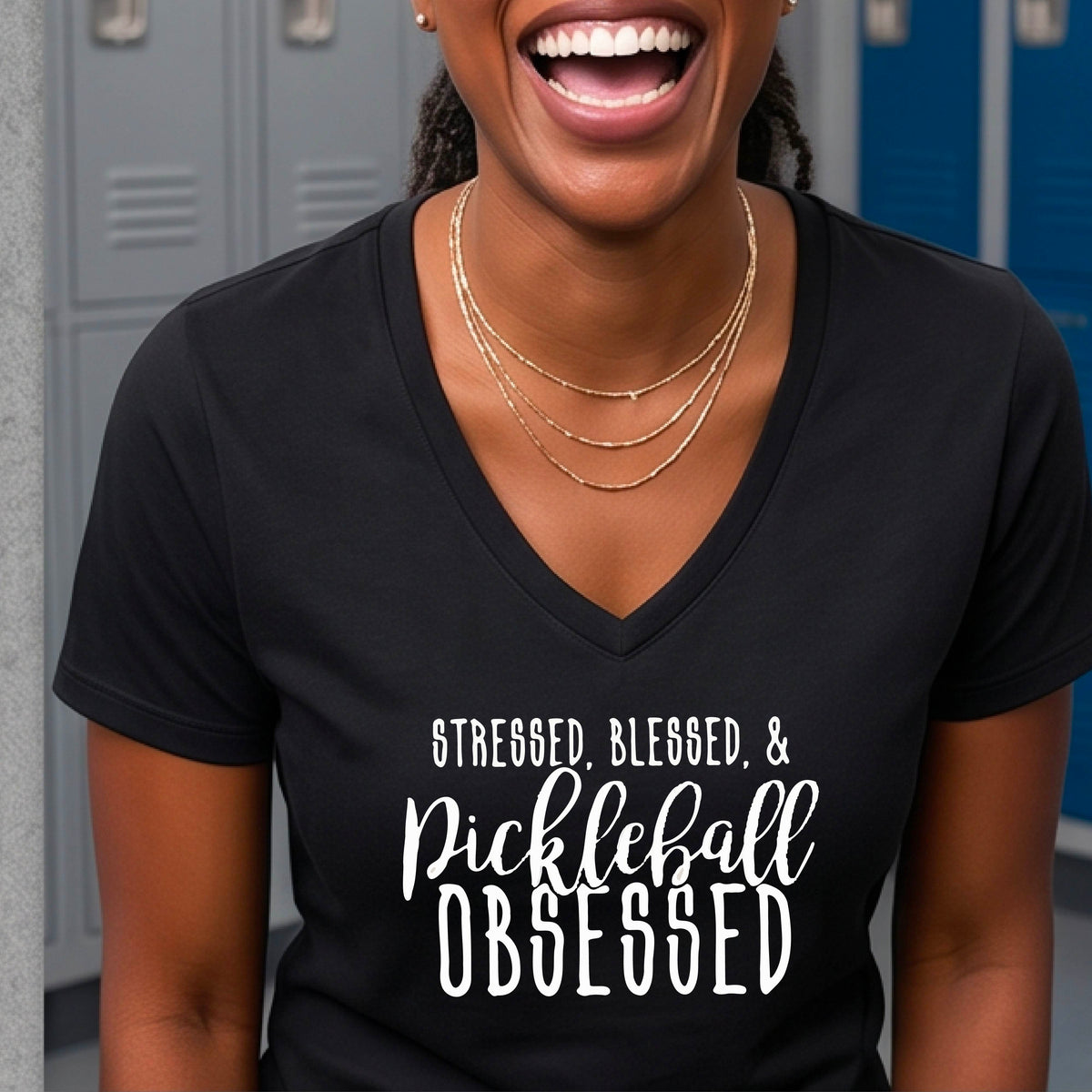 A woman on a pickleball court wearing a black short-sleeve v-neck t-shirt with a white graphic of the words “Stressed, Blessed & Pickleball Obsessed” on the front of the shirt.