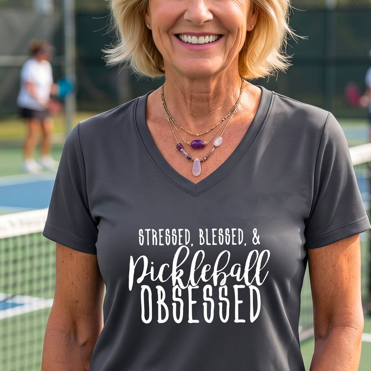 A woman on a pickleball court wearing a dark gray short-sleeve v-neck t-shirt with a white graphic of the words “Stressed, Blessed & Pickleball Obsessed” on the front of the shirt.