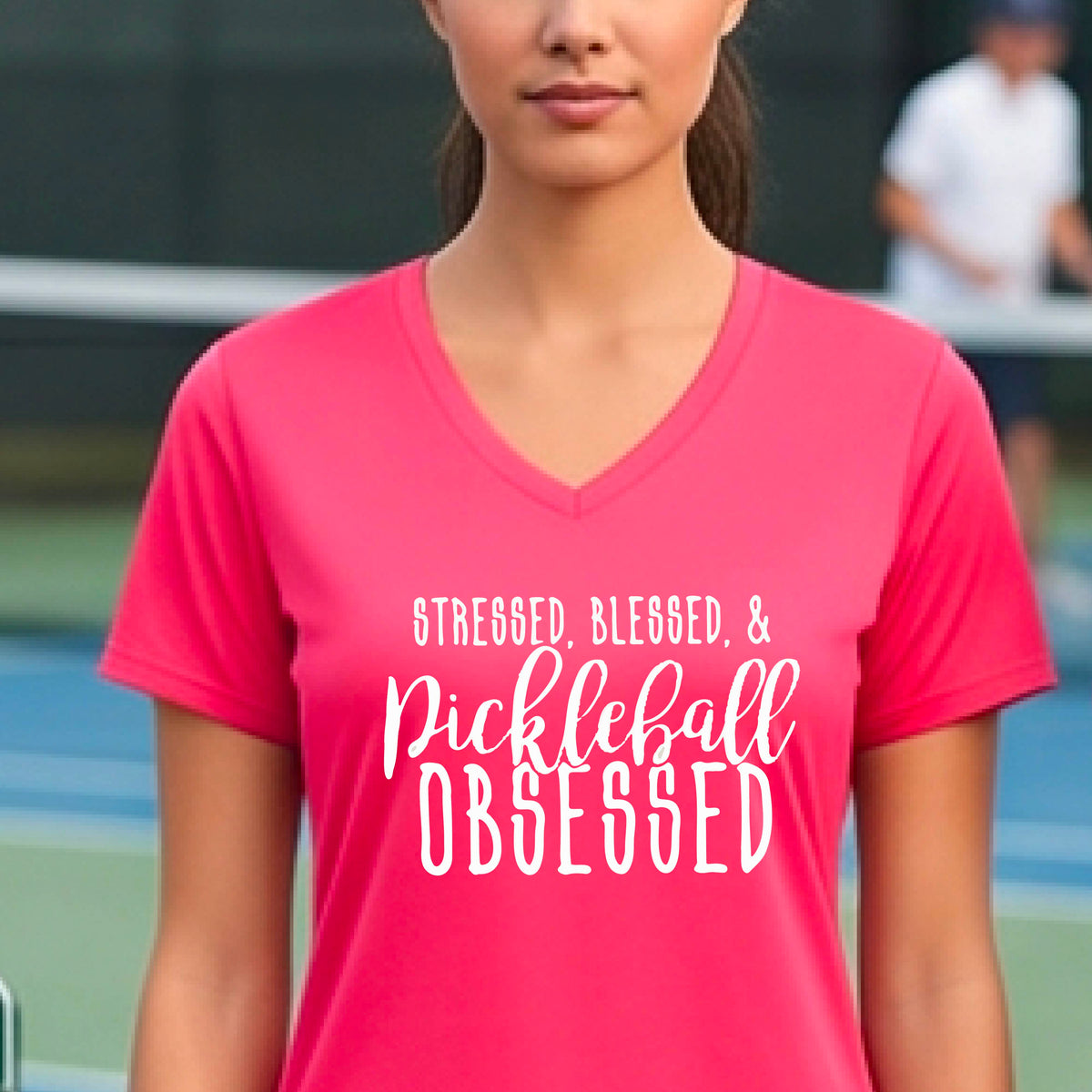 A woman on a pickleball court wearing a coral pink short-sleeve v-neck t-shirt with a white graphic of the words “Stressed, Blessed & Pickleball Obsessed” on the front of the shirt.
