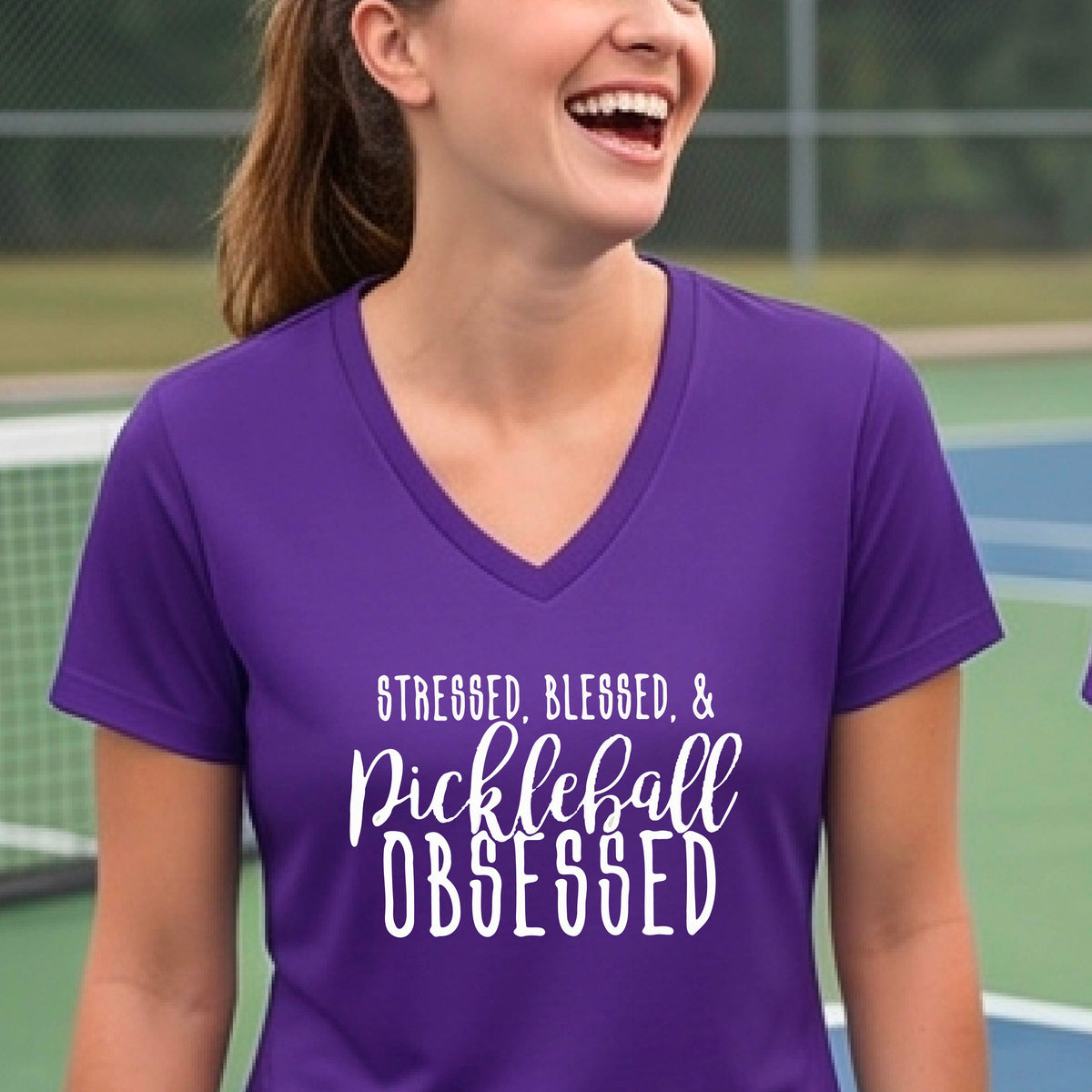 A woman on a pickleball court wearing a purple short-sleeve v-neck t-shirt with a white graphic of the words “Stressed, Blessed & Pickleball Obsessed” on the front of the shirt.