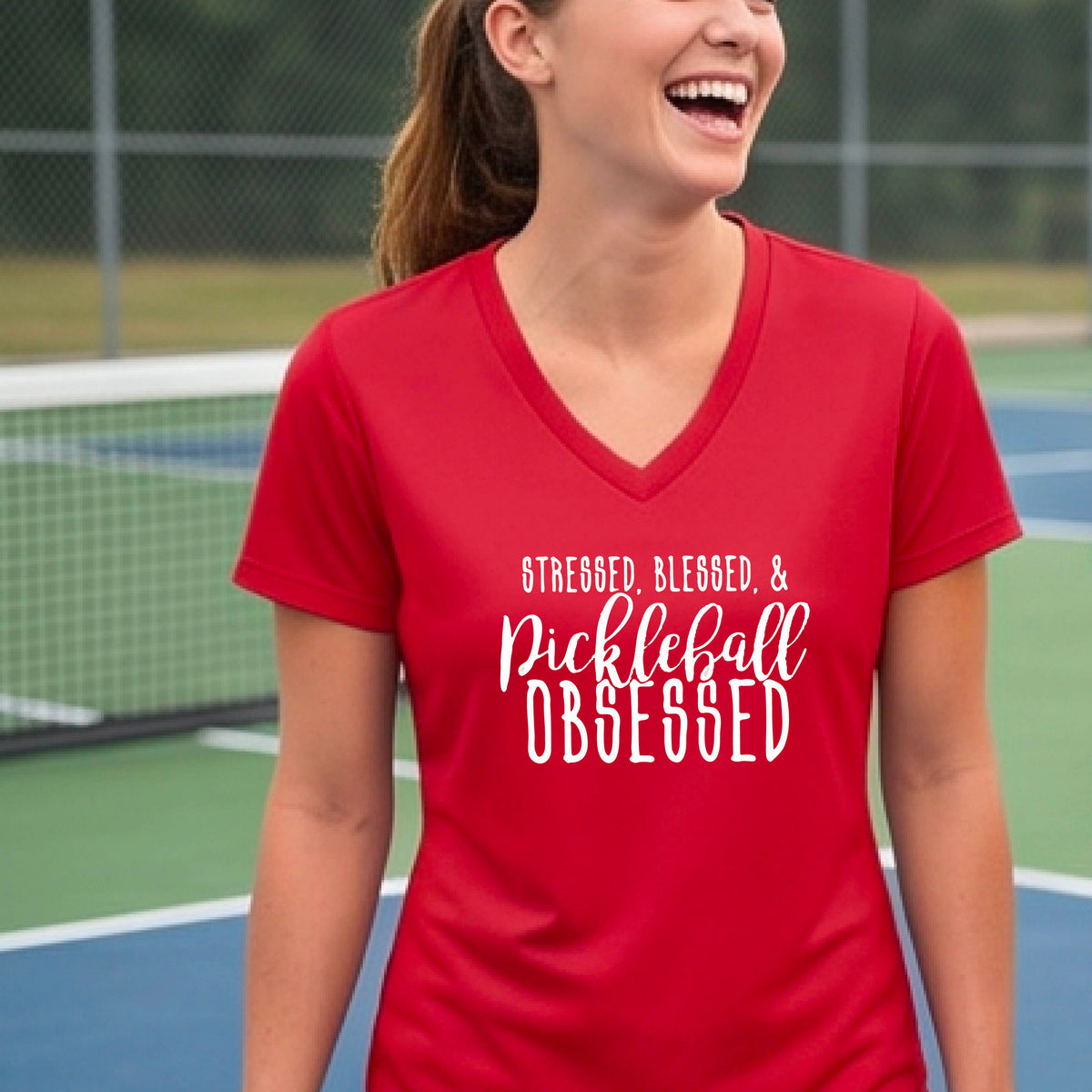 A woman on a pickleball court wearing a red short-sleeve v-neck t-shirt with a white graphic of the words “Stressed, Blessed & Pickleball Obsessed” on the front of the shirt.