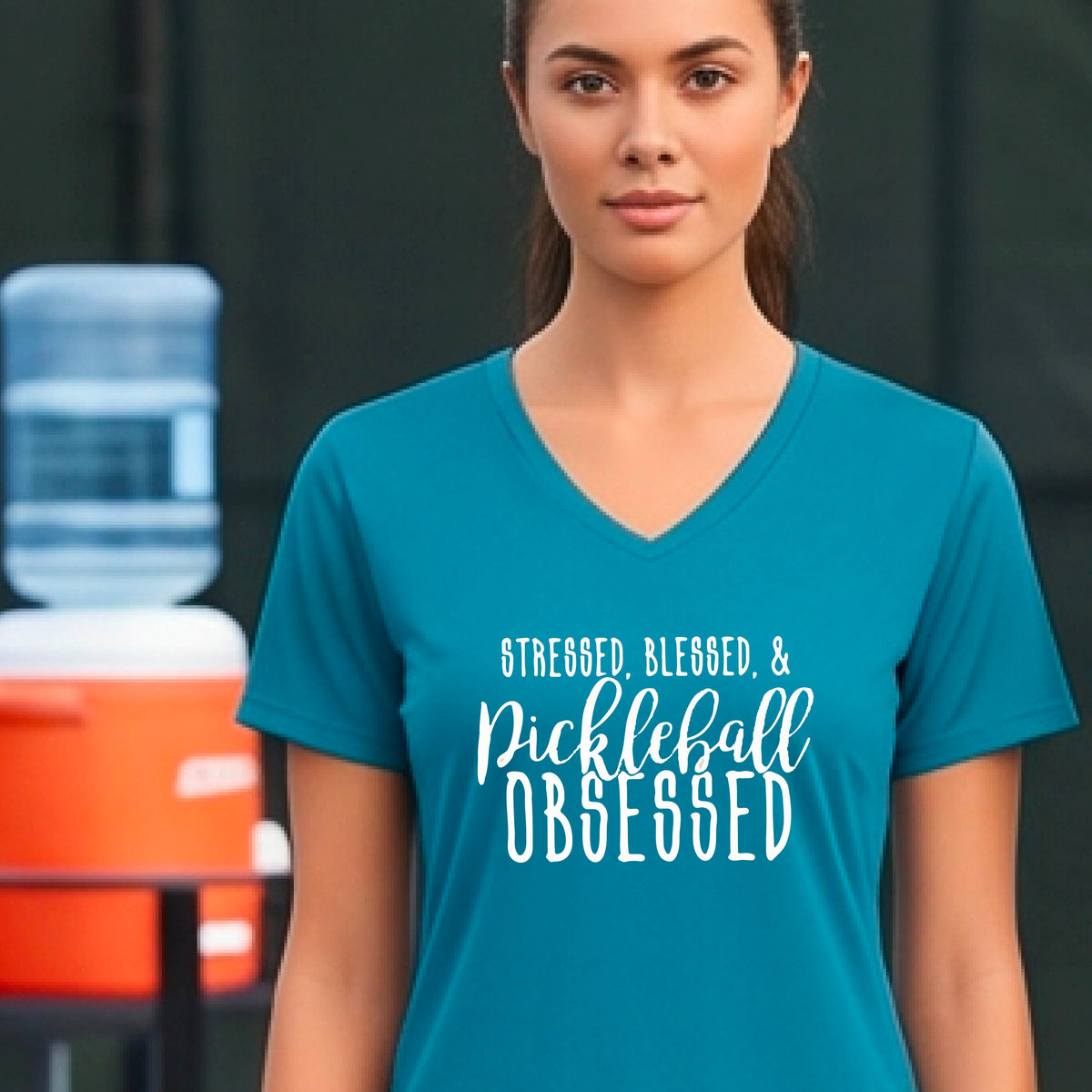 A woman on a pickleball court wearing a teal short-sleeve v-neck t-shirt with a white graphic of the words “Stressed, Blessed & Pickleball Obsessed” on the front of the shirt.