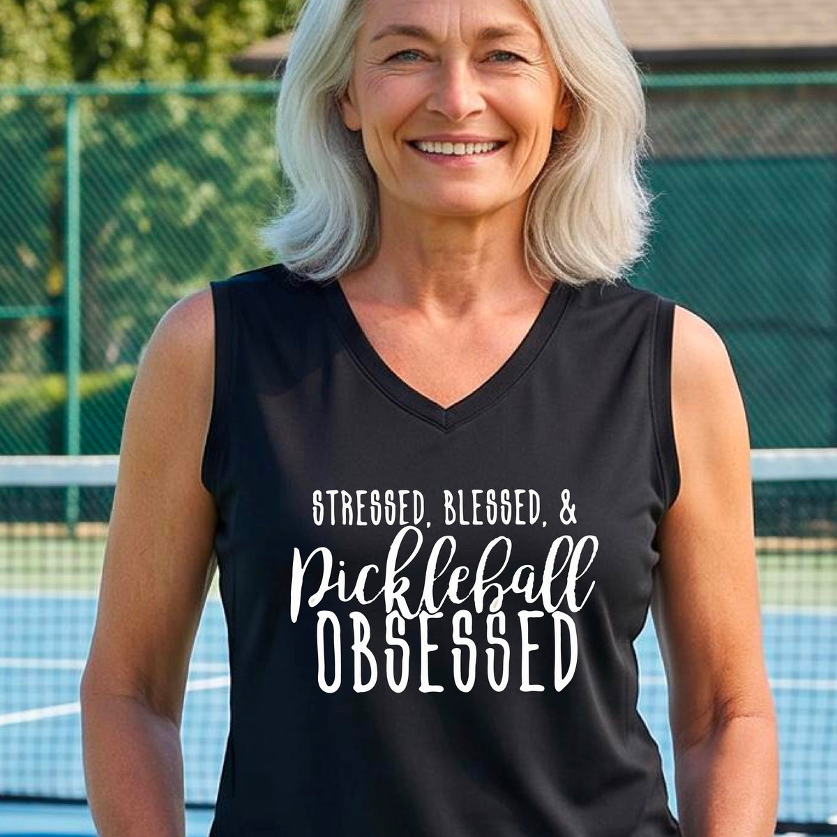 A woman wearing a bright red sleeveless V-neck tank top with white text that reads 'STRESSED, BLESSED, & Pickleball OBSESSED' is standing on a pickleball court.
