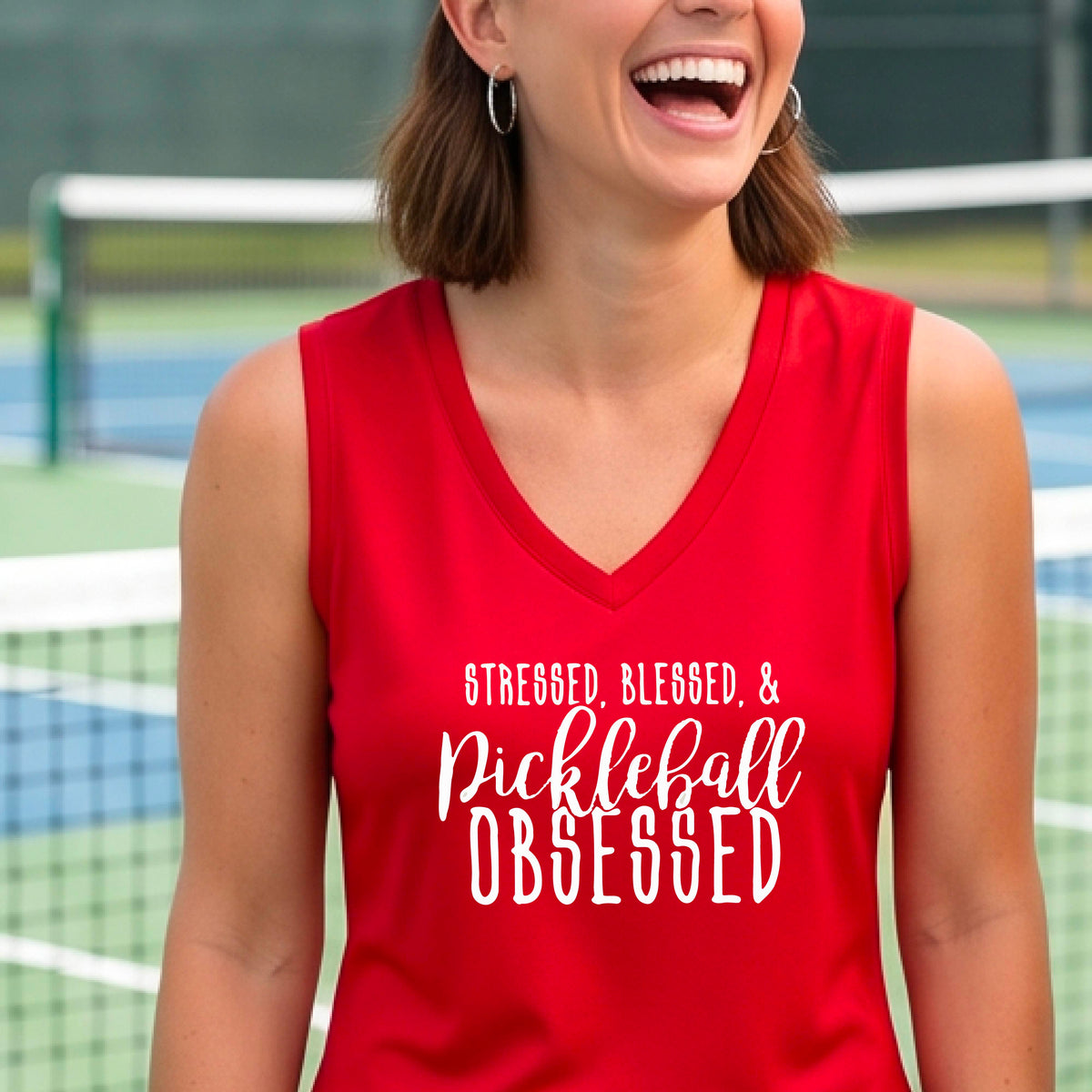 A woman wearing a bright red sleeveless V-neck tank top with white text that reads 'STRESSED, BLESSED, & Pickleball OBSESSED' is standing on a pickleball court.