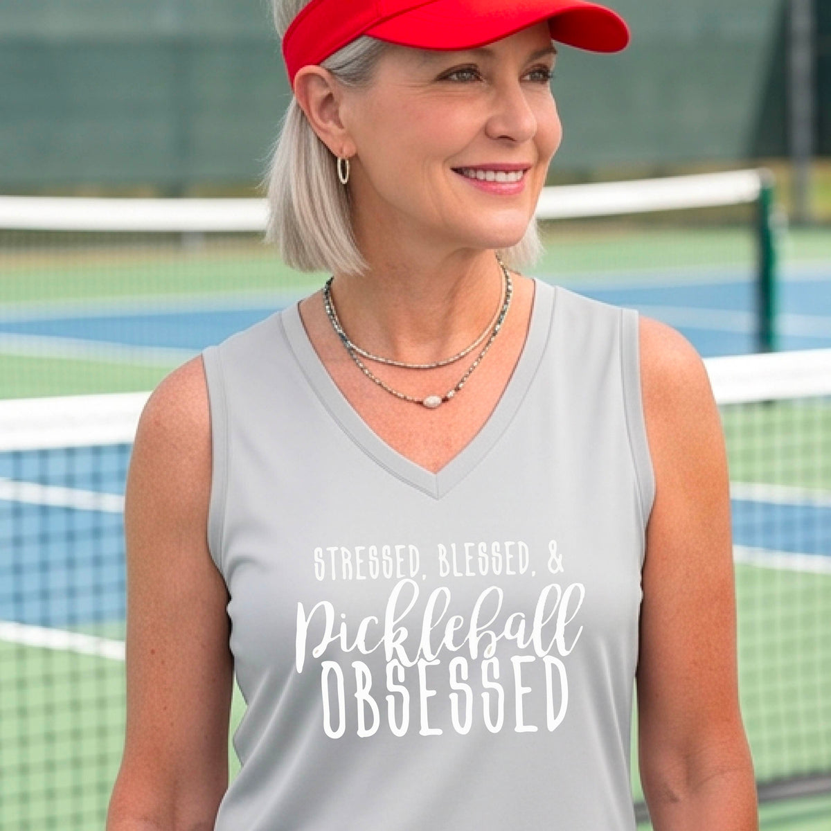 A woman wearing a silver gray sleeveless V-neck tank top with white text that reads 'STRESSED, BLESSED, & Pickleball OBSESSED' is standing on a pickleball court.