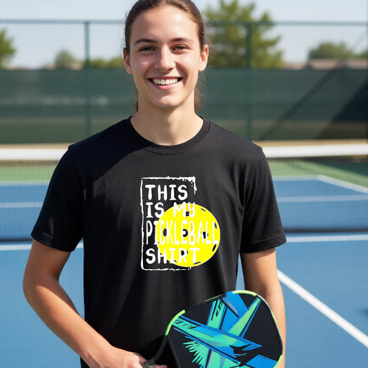 A smiling teenager on a pickleball court wearing a black graphic t-shirt that reads 'THIS IS MY PICKLEBALL SHIRT' in white text over a yellow pickleball,standing in front of a pickleball court.