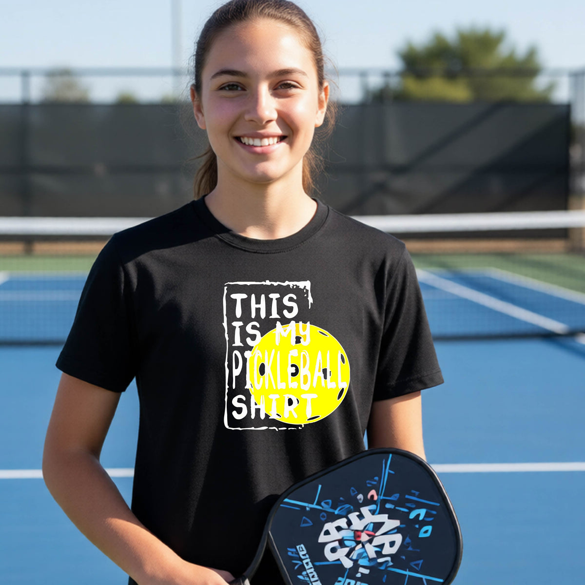 A smiling teenager on a pickleball court wearing a black graphic t-shirt that reads 'THIS IS MY PICKLEBALL SHIRT' in white text over a yellow pickleball,standing in front of a pickleball court.