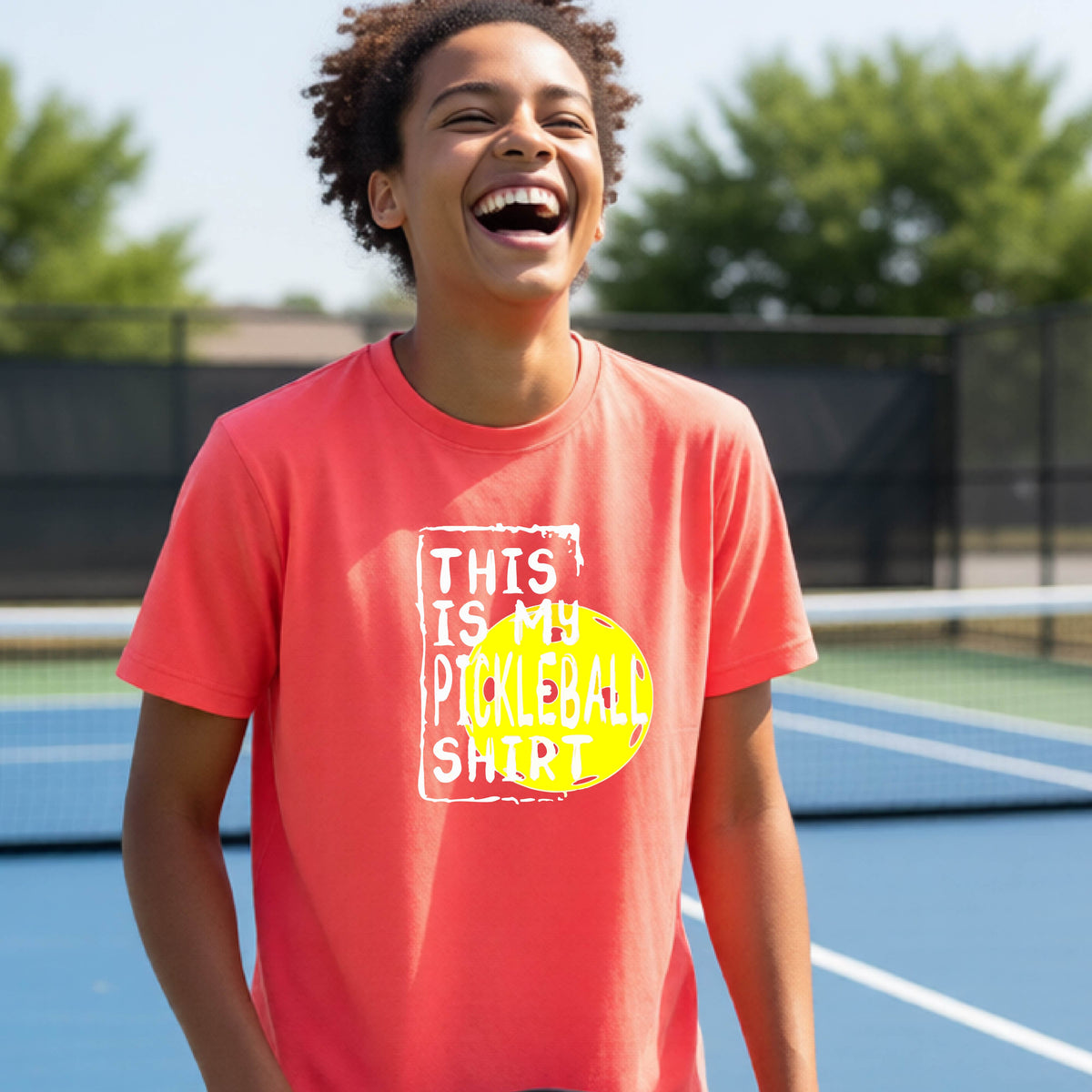 A smiling teenager on a pickleball court wearing a coral pink graphic t-shirt that reads 'THIS IS MY PICKLEBALL SHIRT' in white text over a yellow pickleball,standing in front of a pickleball court.
