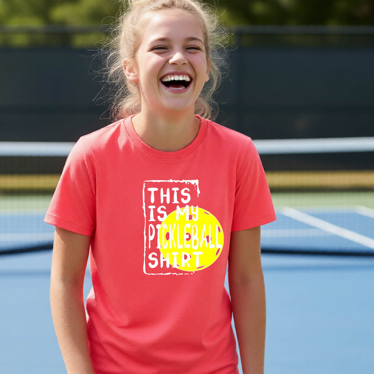 A smiling teenager on a pickleball court wearing a coral pink graphic t-shirt that reads 'THIS IS MY PICKLEBALL SHIRT' in white text over a yellow pickleball,standing in front of a pickleball court.