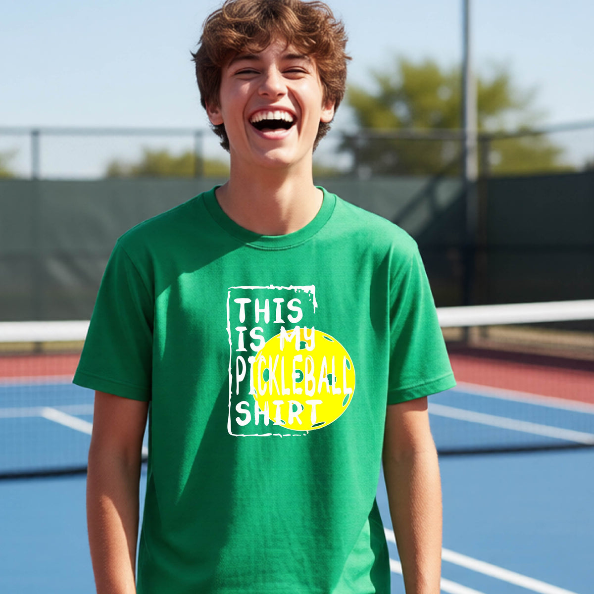 A smiling teenager on a pickleball court wearing a green graphic t-shirt that reads 'THIS IS MY PICKLEBALL SHIRT' in white text over a yellow pickleball,standing in front of a pickleball court.