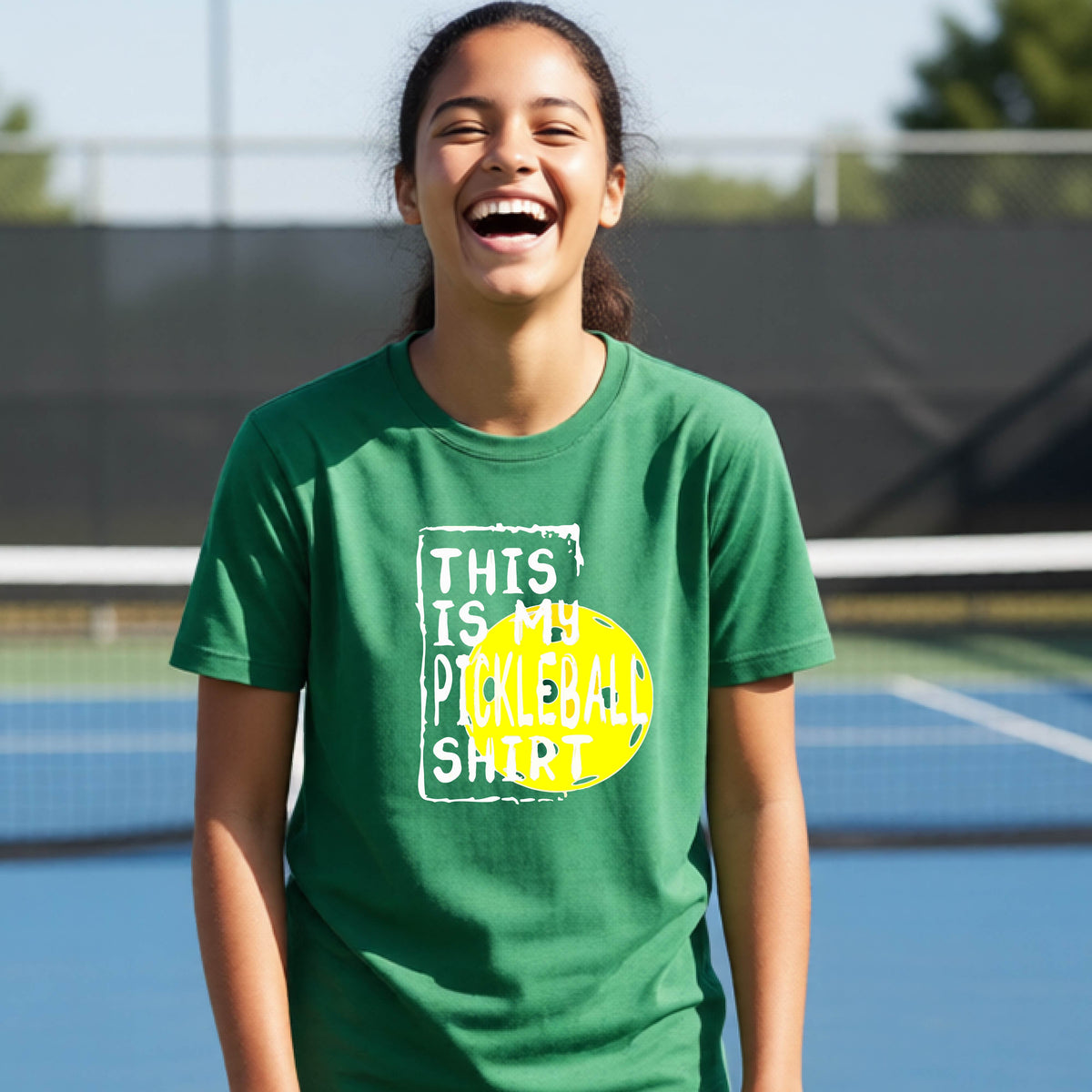 A smiling teenager on a pickleball court wearing a green graphic t-shirt that reads 'THIS IS MY PICKLEBALL SHIRT' in white text over a yellow pickleball,standing in front of a pickleball court.