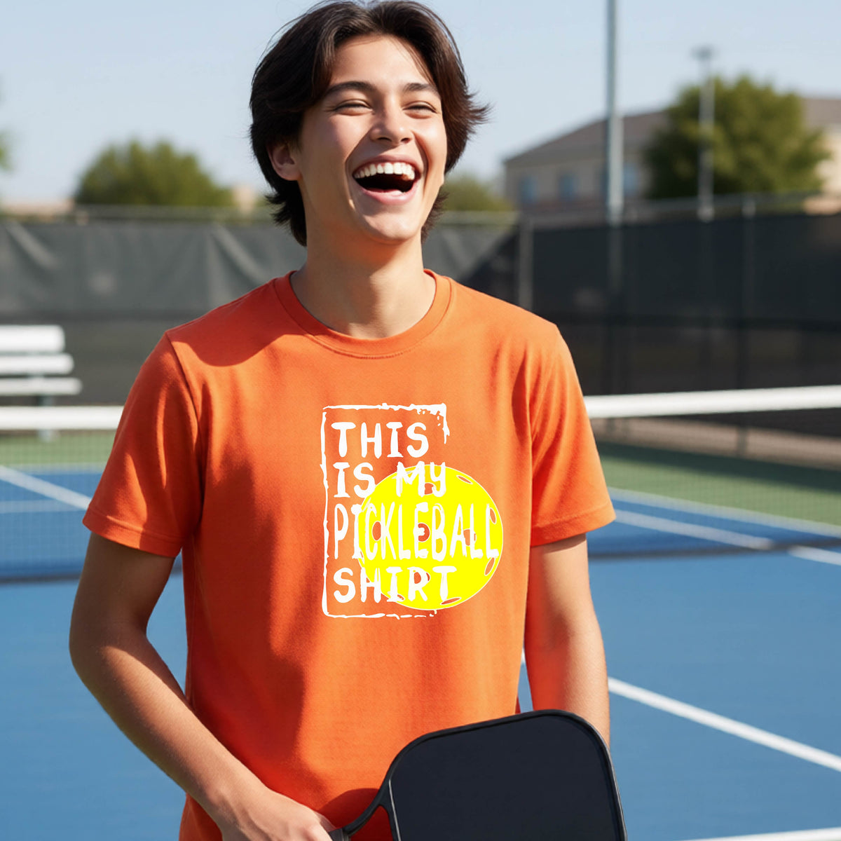 A smiling teenager on a pickleball court wearing an orange graphic t-shirt that reads 'THIS IS MY PICKLEBALL SHIRT' in white text over a yellow pickleball,standing in front of a pickleball court.