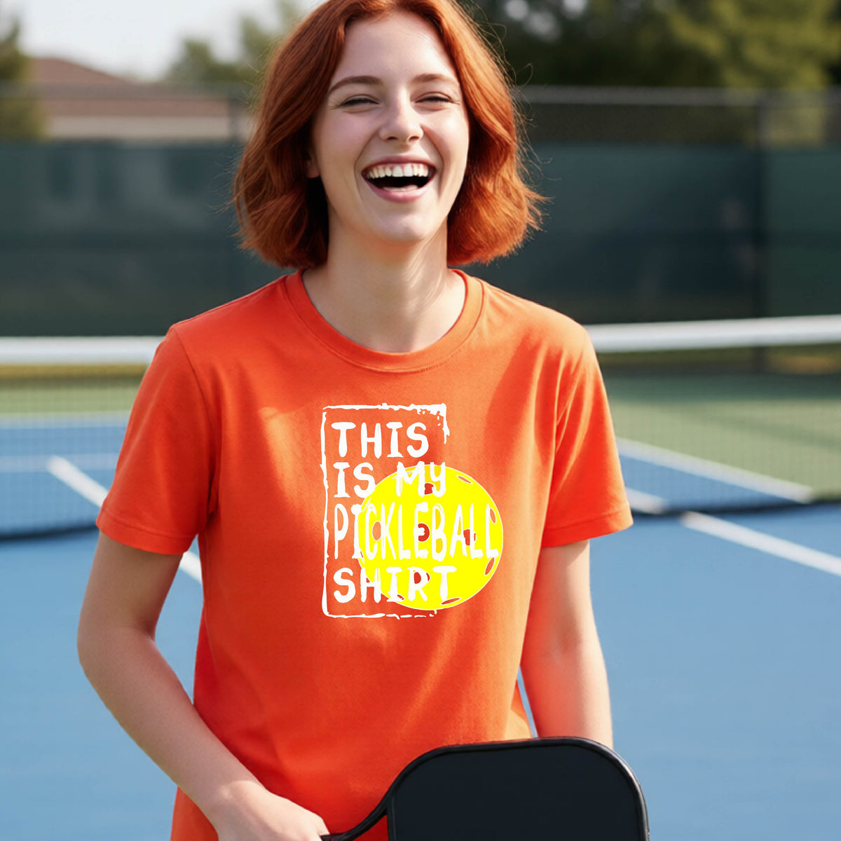 A smiling teenager on a pickleball court wearing an orange graphic t-shirt that reads 'THIS IS MY PICKLEBALL SHIRT' in white text over a yellow pickleball,standing in front of a pickleball court. 
