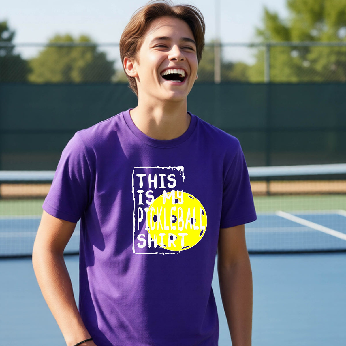 A smiling teenager on a pickleball court wearing a purple graphic t-shirt that reads 'THIS IS MY PICKLEBALL SHIRT' in white text over a yellow pickleball,standing in front of a pickleball court.