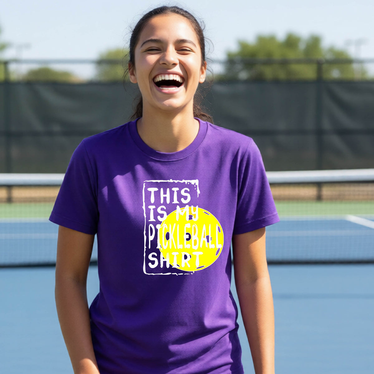 A smiling teenager on a pickleball court wearing a purple graphic t-shirt that reads 'THIS IS MY PICKLEBALL SHIRT' in white text over a yellow pickleball,standing in front of a pickleball court.