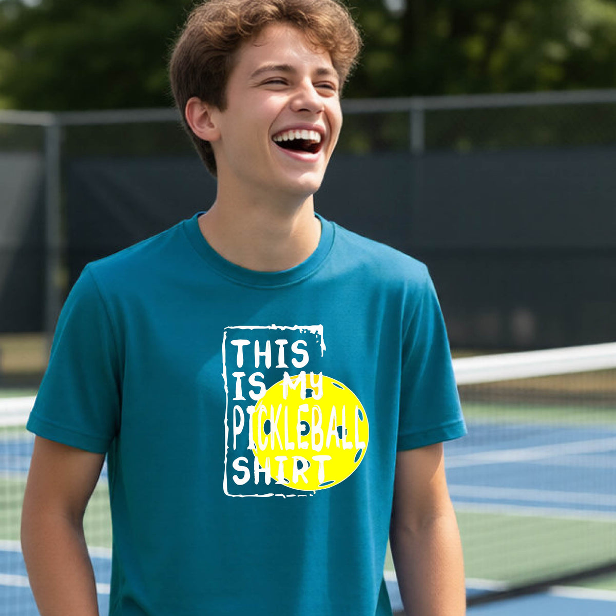A smiling teenager on a pickleball court wearing a teal graphic t-shirt that reads 'THIS IS MY PICKLEBALL SHIRT' in white text over a yellow pickleball,standing in front of a pickleball court.