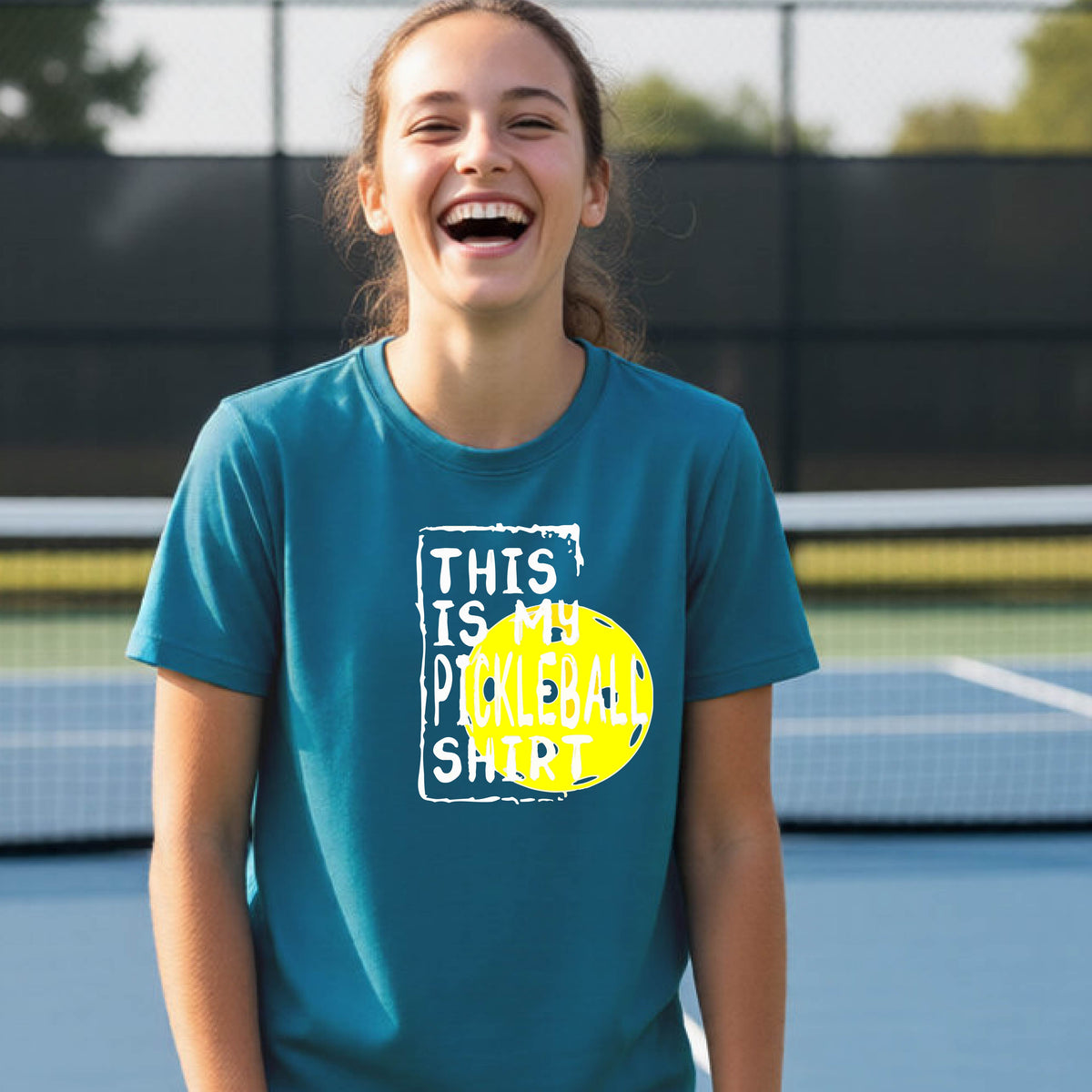 A smiling teenager on a pickleball court wearing a teal  blue graphic t-shirt that reads 'THIS IS MY PICKLEBALL SHIRT' in white text over a yellow pickleball,standing in front of a pickleball court.