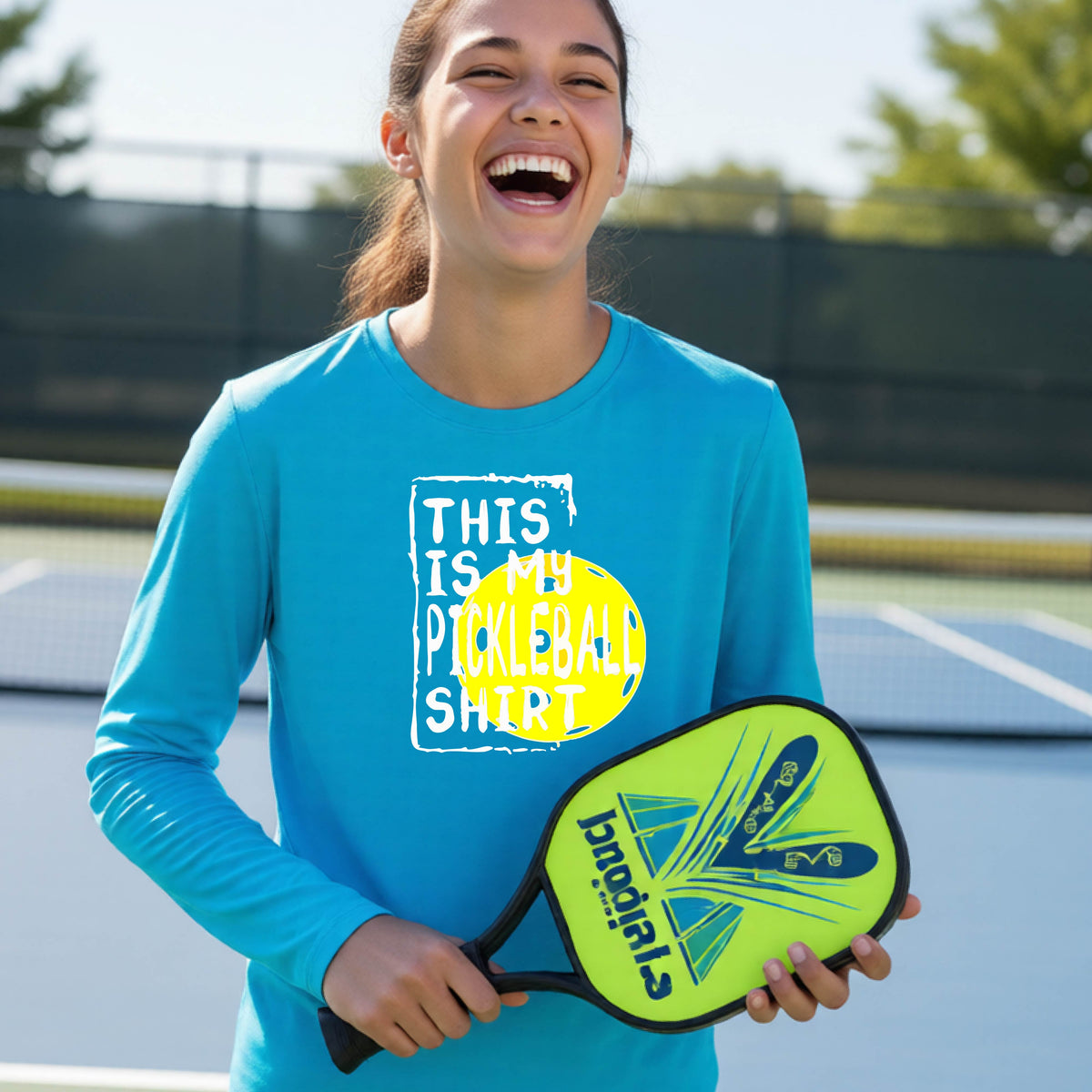 A teenager laughing on an outdoor tennis court wearing a bright blue long-sleeve t-shirt with a graphic that reads 'THIS IS MY PICKLEBALL SHIRT' in white and yellow.