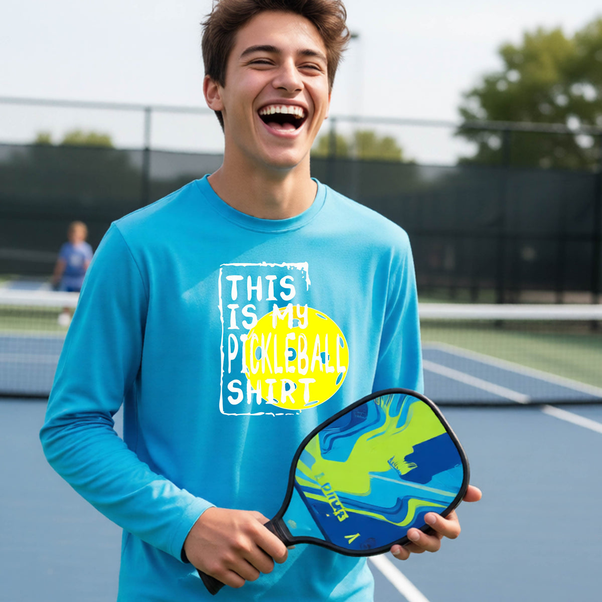 A teenager laughing on an outdoor tennis court wearing a bright blue long-sleeve t-shirt with a graphic that reads 'THIS IS MY PICKLEBALL SHIRT' in white and yellow.