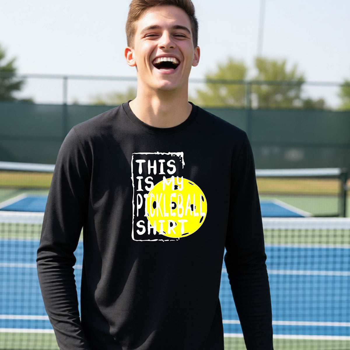 A teenager laughing on an outdoor tennis court wearing a black long-sleeve t-shirt with a graphic that reads 'THIS IS MY PICKLEBALL SHIRT' in white and yellow.