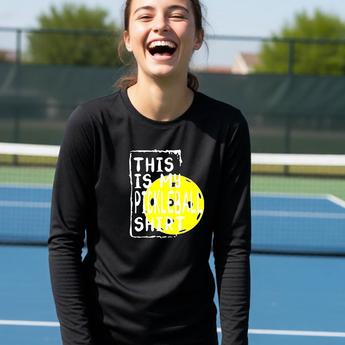 A teenager laughing on an outdoor tennis court wearing a black long-sleeve t-shirt with a graphic that reads 'THIS IS MY PICKLEBALL SHIRT' in white and yellow.