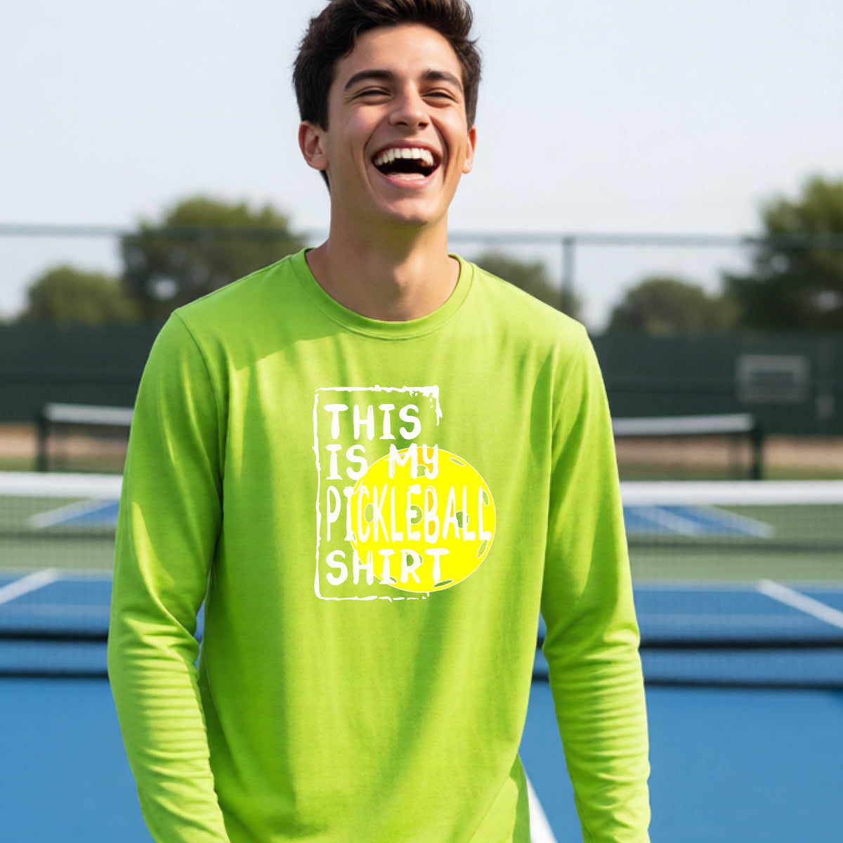 A teenager laughing on an outdoor tennis court wearing a lime green long-sleeve t-shirt with a graphic that reads 'THIS IS MY PICKLEBALL SHIRT' in white and yellow.