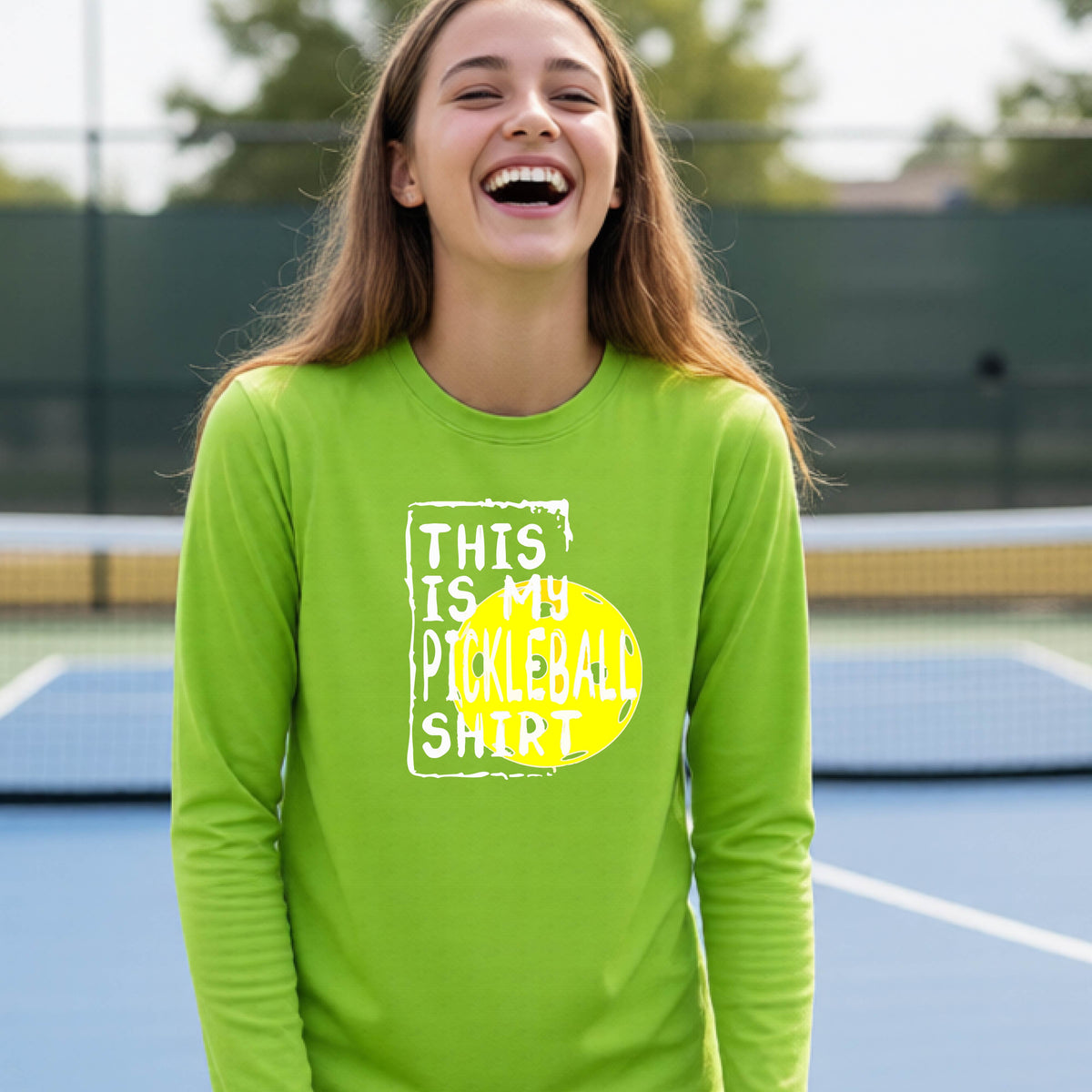 A teenager laughing on an outdoor tennis court wearing a lime green long-sleeve t-shirt with a graphic that reads 'THIS IS MY PICKLEBALL SHIRT' in white and yellow.