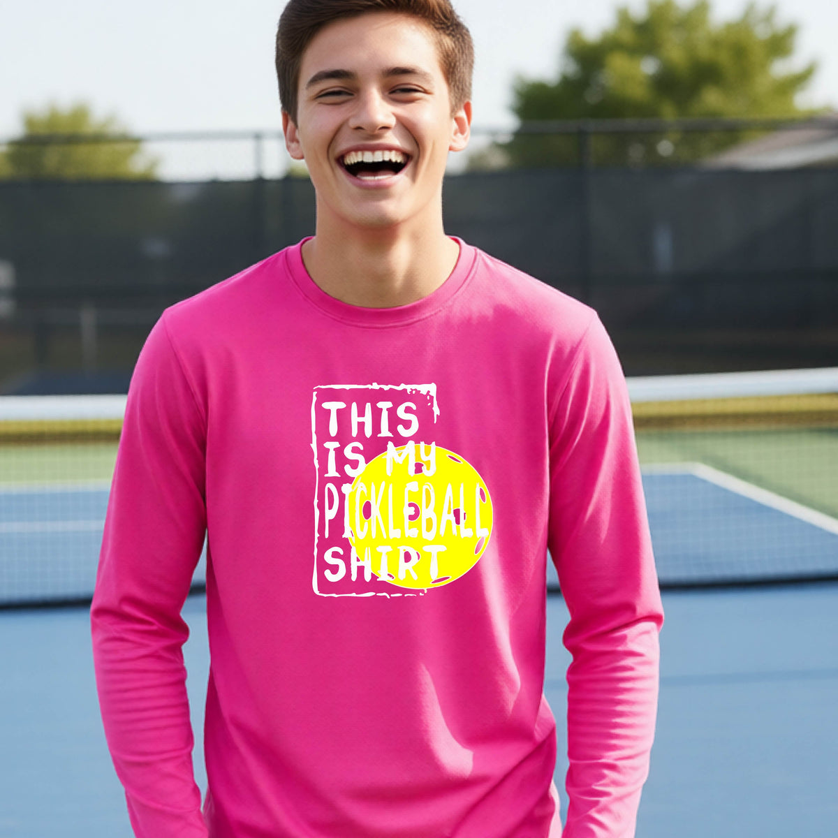 A teenager laughing on an outdoor tennis court wearing a pink long-sleeve t-shirt with a graphic that reads 'THIS IS MY PICKLEBALL SHIRT' in white and yellow.