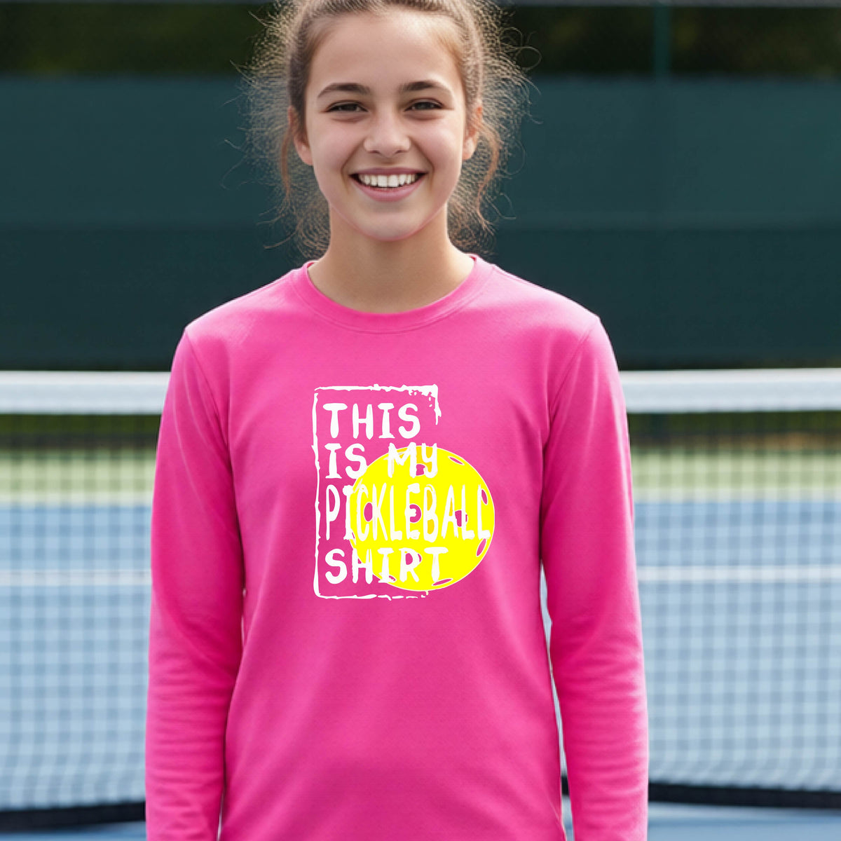 A teenager laughing on an outdoor tennis court wearing a pink long-sleeve t-shirt with a graphic that reads 'THIS IS MY PICKLEBALL SHIRT' in white and yellow.