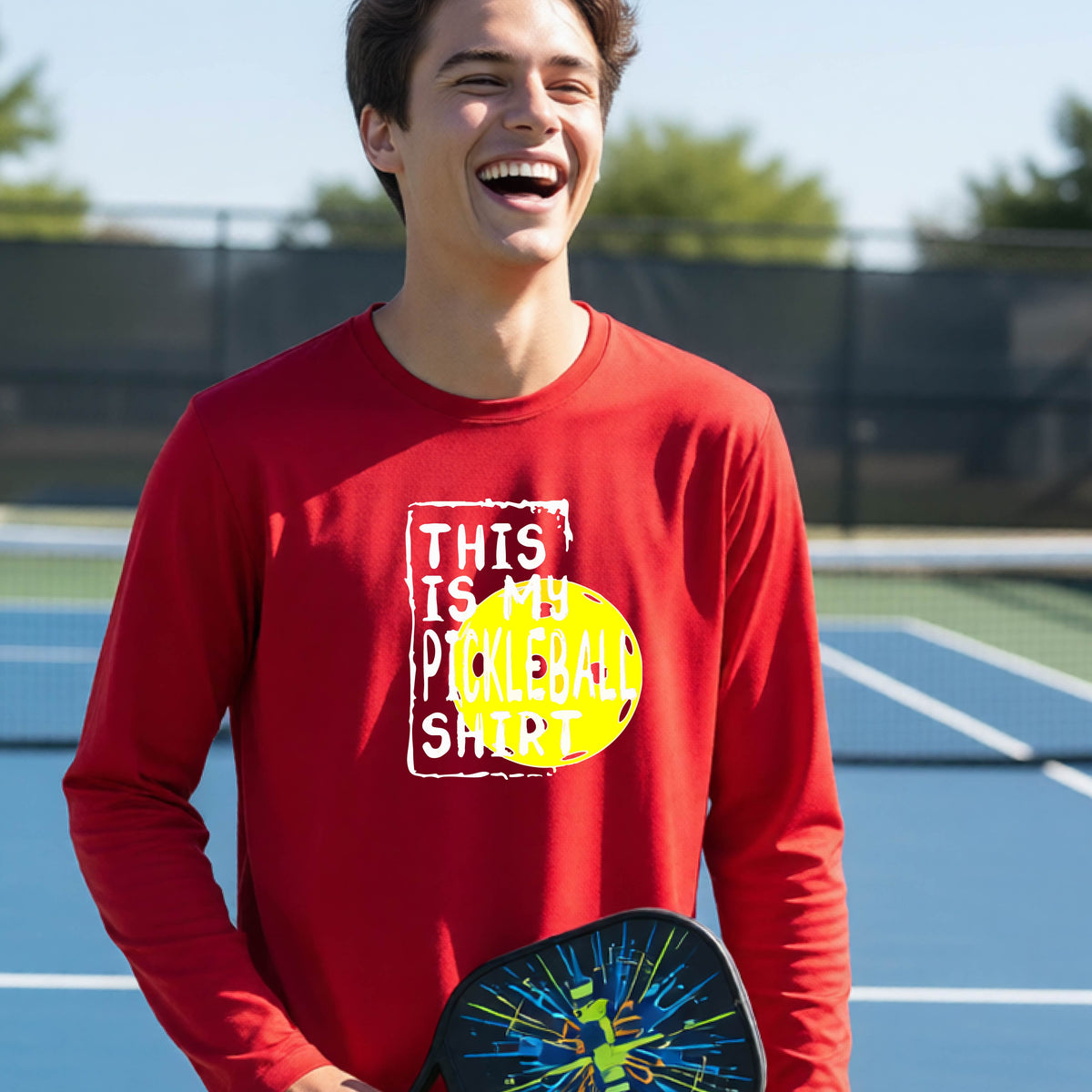 A teenager laughing on an outdoor tennis court wearing a red long-sleeve t-shirt with a graphic that reads 'THIS IS MY PICKLEBALL SHIRT' in white and yellow.