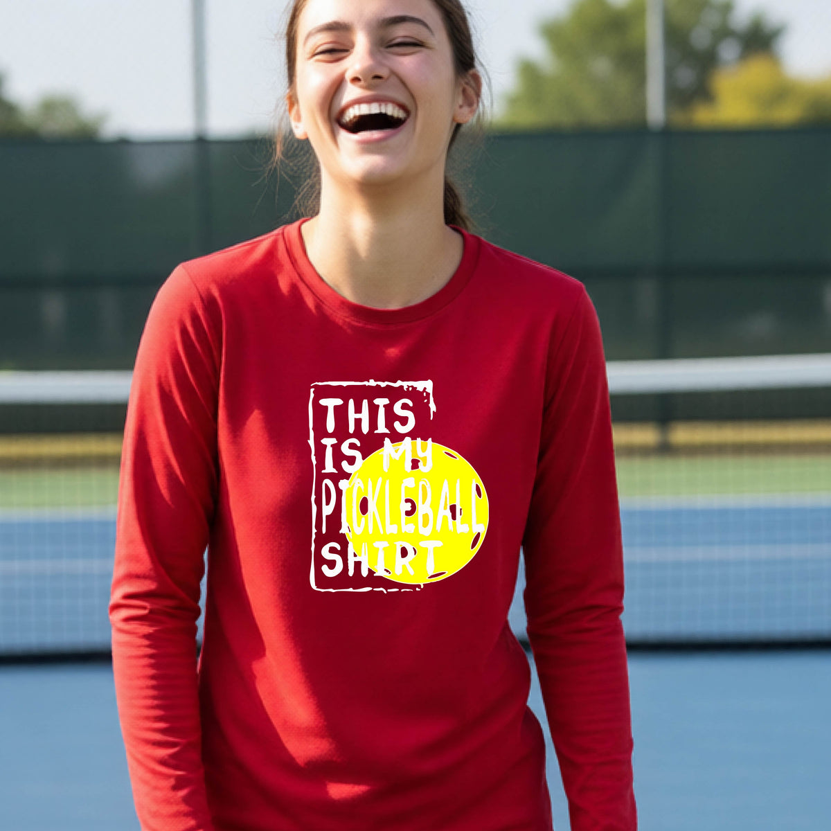 A teenager laughing on an outdoor tennis court wearing a red long-sleeve t-shirt with a graphic that reads 'THIS IS MY PICKLEBALL SHIRT' in white and yellow.