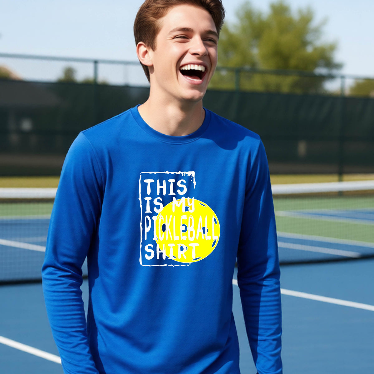 A teenager laughing on an outdoor tennis court wearing a bright blue long-sleeve t-shirt with a graphic that reads 'THIS IS MY PICKLEBALL SHIRT' in white and yellow.