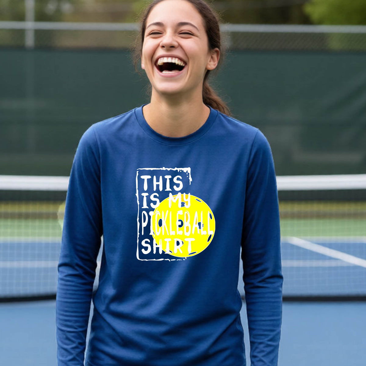 A teenager laughing on an outdoor tennis court wearing a royal blue long-sleeve t-shirt with a graphic that reads 'THIS IS MY PICKLEBALL SHIRT' in white and yellow.