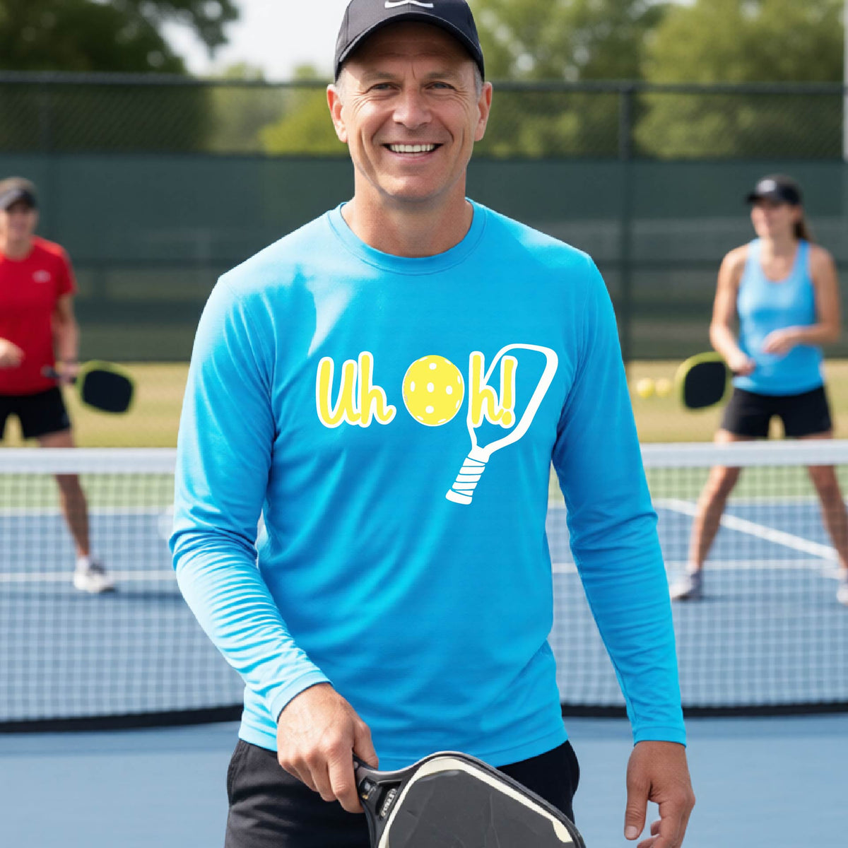 A man on a pickleball court wearing a bright blue long-sleeve t-shirt with a white and yellow graphic of the words “Uh Oh!” with a yellow pickleball replacing the letter O on the front of the shirt, over the top of a pickleball paddle.