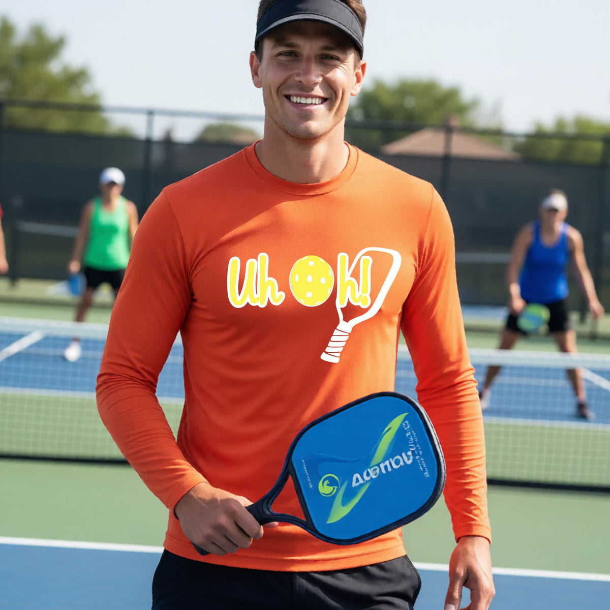 A man on a pickleball court wearing an orange long-sleeve t-shirt with a white and yellow graphic of the words “Uh Oh!” with a yellow pickleball replacing the letter O on the front of the shirt, over the top of a pickleball paddle.