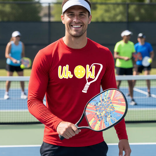 A man on a pickleball court wearing a red long-sleeve t-shirt with a white and yellow graphic of the words “Uh Oh!” with a yellow pickleball replacing the letter O on the front of the shirt, over the top of a pickleball paddle.