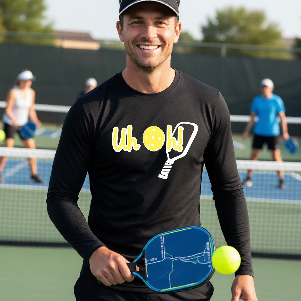 A man on a pickleball court wearing a black long-sleeve t-shirt with a white and yellow graphic of the words “Uh Oh!” with a yellow pickleball replacing the letter O on the front of the shirt, over the top of a pickleball paddle.
