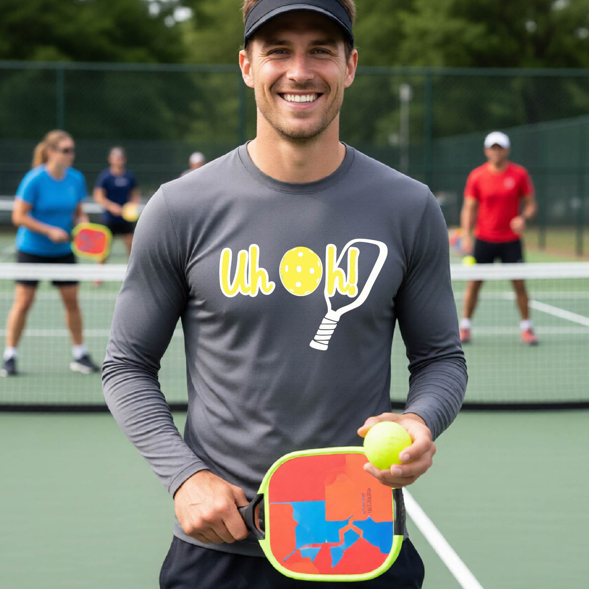 A man on a pickleball court wearing a gray long-sleeve t-shirt with a white and yellow graphic of the words “Uh Oh!” with a yellow pickleball replacing the letter O on the front of the shirt, over the top of a pickleball paddle.