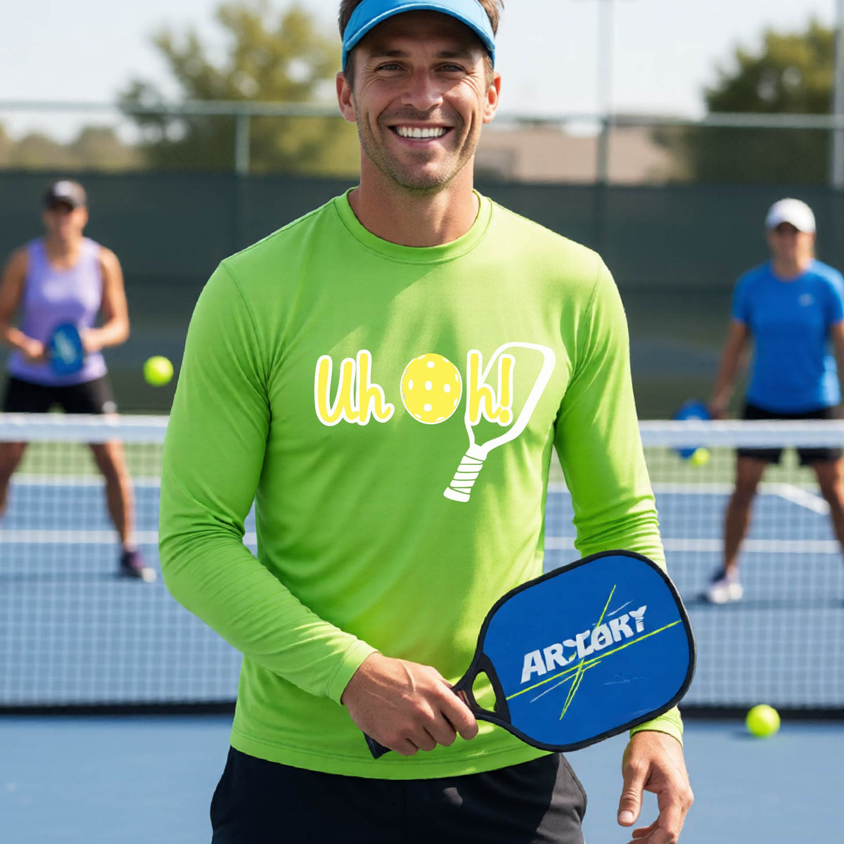 A man on a pickleball court wearing a lime green long-sleeve t-shirt with a white and yellow graphic of the words “Uh Oh!” with a yellow pickleball replacing the letter O on the front of the shirt, over the top of a pickleball paddle.