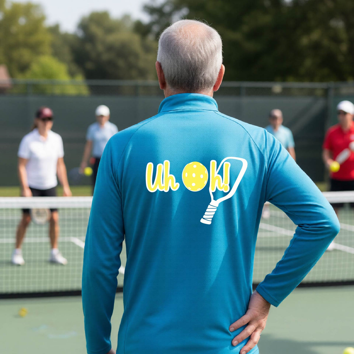 A man seen from behind on a pickleball court wearing a bright blue quarter-zip shirt with a white and yellow graphic of the words “Uh Oh!” with a yellow pickleball replacing the letter O, over the top of a pickleball paddle, on the back of the shirt.