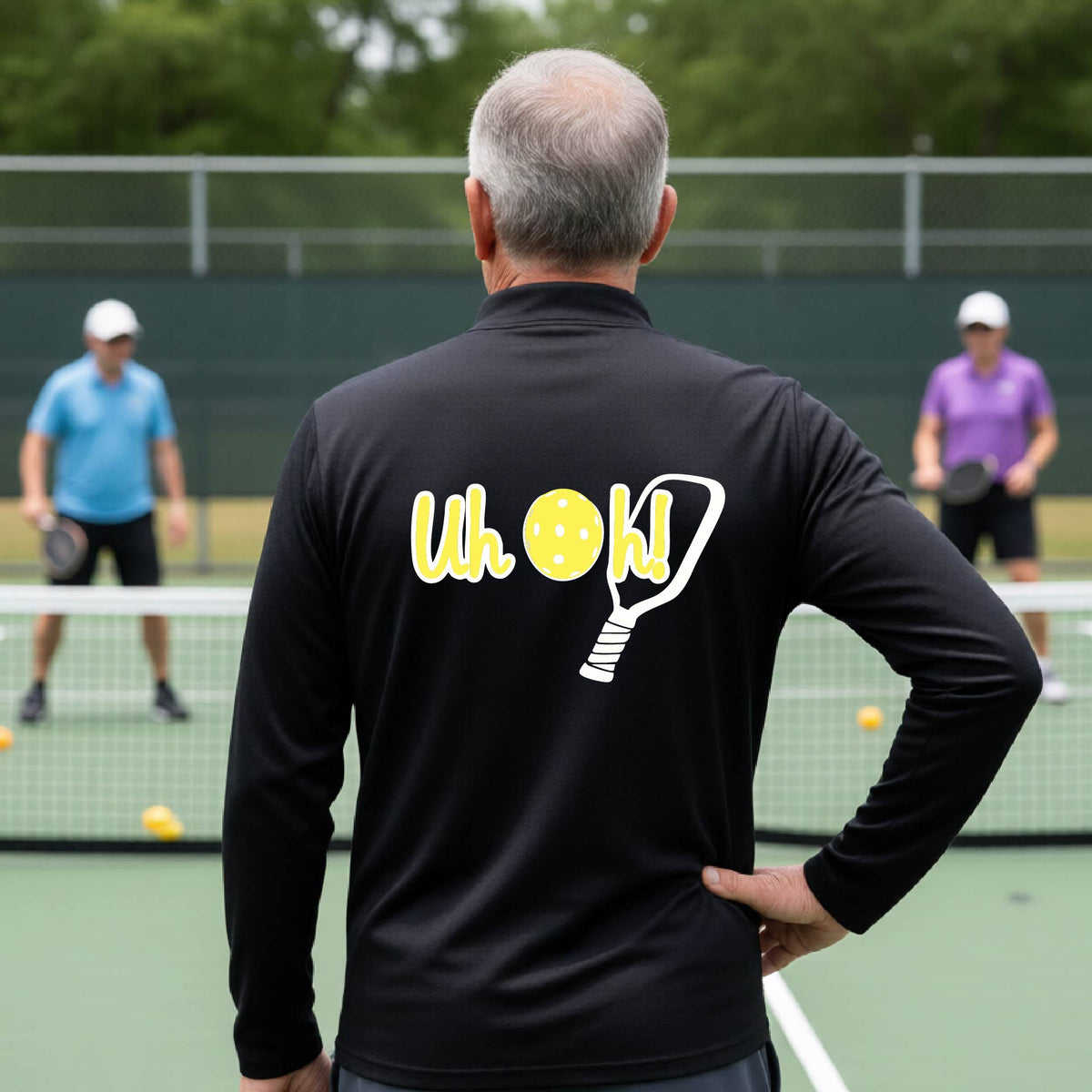 A man seen from behind on a pickleball court wearing a black quarter-zip shirt with a white and yellow graphic of the words “Uh Oh!” with a yellow pickleball replacing the letter O, over the top of a pickleball paddle, on the back of the shirt.