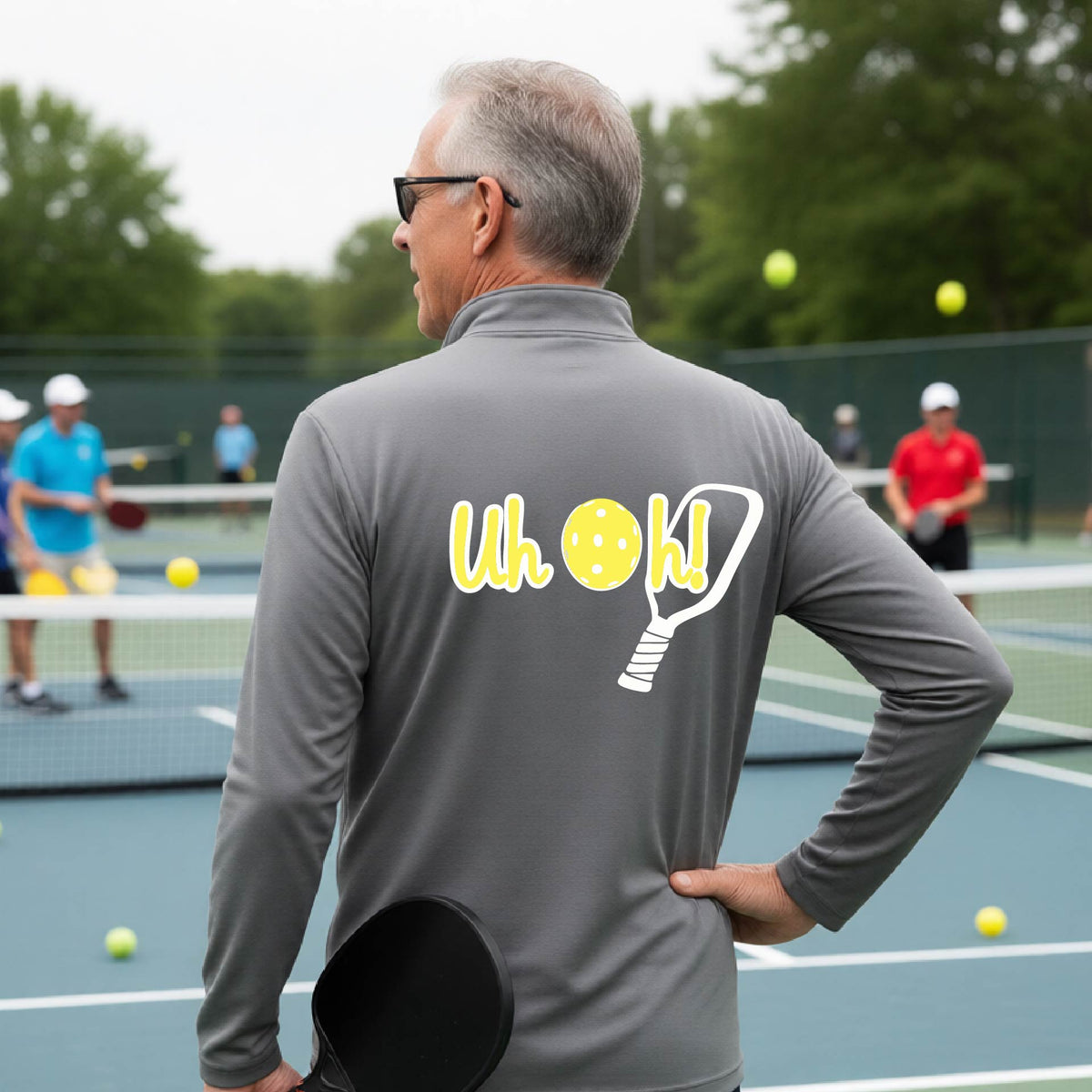 A man seen from behind on a pickleball court wearing a gray quarter-zip shirt with a white and yellow graphic of the words “Uh Oh!” with a yellow pickleball replacing the letter O, over the top of a pickleball paddle, on the back of the shirt.