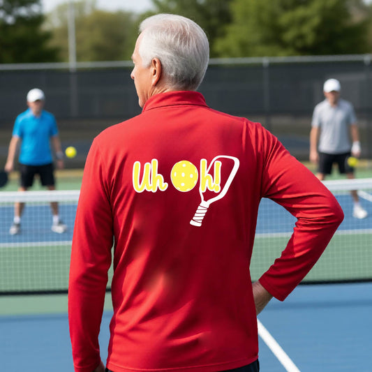 A man seen from behind on a pickleball court wearing a red quarter-zip shirt with a white and yellow graphic of the words “Uh Oh!” with a yellow pickleball replacing the letter O, over the top of a pickleball paddle, on the back of the shirt.